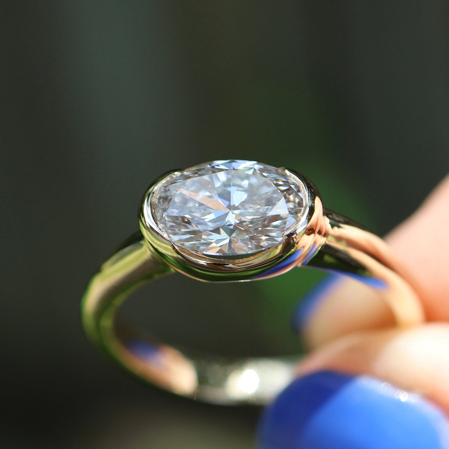 Gold ring with a large diamond held by a hand with blue nail polish against a blurred background