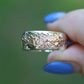 Silver ring with a textured design held by a hand with blue nail polish against a green background