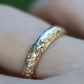Gold ring with intricate design on a finger against a blurred green background