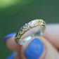 Gold and diamond ring held by a hand with blue nail polish against a blurred green background