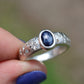 Silver ring with a blue stone held by a hand with purple nail polish against a blurred natural background.